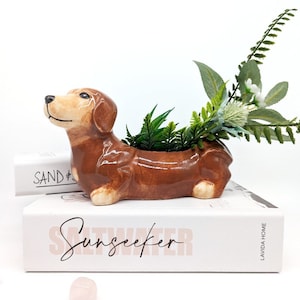 May include: A brown ceramic dachshund planter with green and white artificial plants inside. The planter is sitting on top of a book titled "Saltwater Sunseeker" by Lavida Home.
