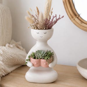May include: A white ceramic vase shaped like a seated figure with a closed-eye face. The figure holds a small, pink, heart-shaped planter containing green succulents. The top of the head is filled with a bouquet of dried grasses and pink flowers.