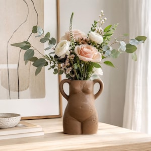 May include: A matte brown ceramic vase shaped like a human torso with two handles, filled with a floral arrangement of light pink roses, white ranunculus, small white filler flowers, and green eucalyptus branches with long, slender stems.