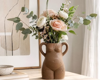 May include: A matte brown ceramic vase shaped like a human torso with two handles, filled with a floral arrangement of light pink roses, white ranunculus, small white filler flowers, and green eucalyptus branches with long, slender stems.