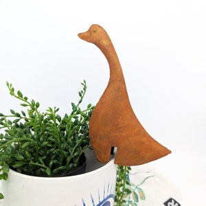 Peut inclure: Une figurine de canard en métal brun rouillé avec un long cou et un corps large. Le canard est perché sur le bord d'un pot en céramique blanc avec une plante succulente verte.