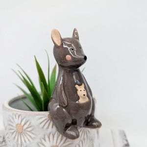 May include: A grey ceramic kangaroo figurine with a joey in its pouch. The kangaroo is sitting on the edge of a white planter with a daisy pattern.