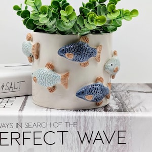 May include: A white ceramic planter with blue and light blue fish decorations and brown fins. The planter is filled with green faux foliage. The planter is sitting on top of a book with the text "ALWAYS IN SEARCH OF THE PERFECT WAVE".