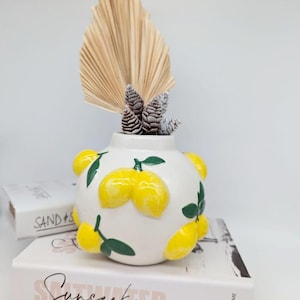 May include: White ceramic vase with a round shape and a lemon design. The vase is decorated with yellow lemons and green leaves.