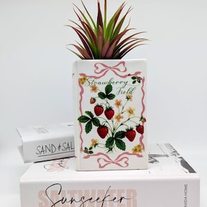 Könnte beinhalten: Ein weißer Keramik-Blumentopf mit einem "Strawberry Field"-Design, das rote Erdbeeren, grüne Blätter und gelbe Blüten zeigt. Der Topf enthält eine Sukkulente mit grünen und burgunderfarbenen Blättern.