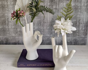 May include: Two white ceramic hand-shaped vases, one with a peace sign gesture and the other with a rock and roll hand gesture, holding pink and white flowers. The vases are on a purple book with the title "Antony and Cleopatra" by Adrian Goldsworthy.