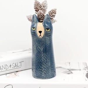 Peut inclure: Un vase en céramique bleu en forme de chat avec un visage blanc et beige et des yeux jaunes. Le vase est décoré d'un motif texturé qui ressemble à de la fourrure. Le vase est rempli de fleurs séchées blanches et grises.