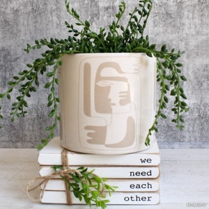 May include: White ceramic planter with a beige abstract face design, filled with a green succulent plant. The planter is sitting on a stack of white books with the text "we need each other" written on them.