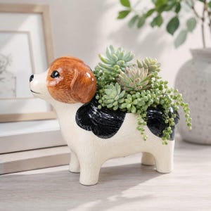 May include: A ceramic dog-shaped planter with a cream-coloured body, black spots, and a brown head. The planter is filled with a variety of green succulents, including a trailing string of pearls plant, creating a decorative botanical display.