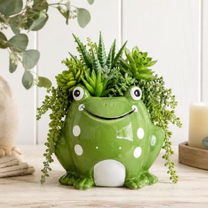 May include: A glossy green ceramic frog planter featuring a wide smiling mouth, two large eyes, and white circular spots on its sides. The frog is filled with a variety of green artificial succulents and trailing foliage that spill over the edges.