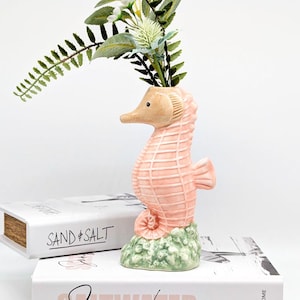 May include: A ceramic seahorse vase in a light pink hue, with a beige head and a green base. The vase holds greenery and white flowers. The words "SAND & SALT" are visible on a book in the background.