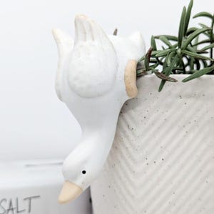 May include: White ceramic duck planter with a small plant growing out of its back. The duck is positioned as if it is hanging upside down from the edge of a white planter with a chevron pattern.