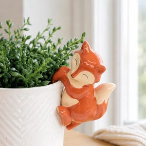 May include: A glossy ceramic fox figurine in shades of orange and cream, designed to hang onto the rim of a white planter. The fox has a smooth finish, pointed ears, and a curved tail, with its front paws hooked over the edge of the container.