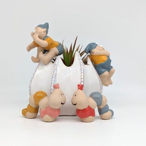Può includere: Un vaso di ceramica bianca a forma di fiore con tre piccole figurine di ceramica. Due delle figurine indossano abiti gialli e blu e la terza indossa un cappello blu e una camicia gialla. Il vaso ha una piccola pianta verde che cresce dalla cima.
