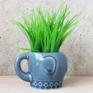 Può includere: Un vaso di ceramica blu a forma di elefante con manico. Il vaso è riempito con erba artificiale verde.