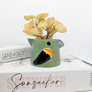 Puede incluir: Una maceta de cerámica verde con forma de pájaro con un diseño negro, naranja y amarillo, que contiene una pequeña planta con hojas amarillas. La maceta está colocada sobre un libro blanco con la palabra "Sunseeker" en cursiva.