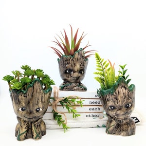 May include: Three ceramic Groot-inspired planters, each holding different faux succulents. The planters are brown with a wood-grain texture and green trim. The planters have smiling faces and are arranged on a white surface with a stack of books.