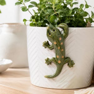 May include: A white ceramic plant pot featuring a subtle chevron texture and a three-dimensional green lizard figurine attached to the side. The lizard has a glossy finish with small orange spots along its back and is positioned as if climbing.