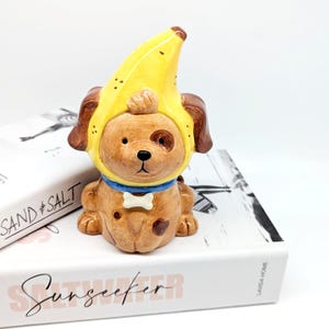 Pode incluir: Estatueta de cão de cerâmica usando um chapéu de banana e uma coleira azul com uma etiqueta em forma de osso. O cão é marrom com manchas e orelhas marrons escuras. O chapéu de banana é amarelo com detalhes marrons. A estatueta está em um livro branco.