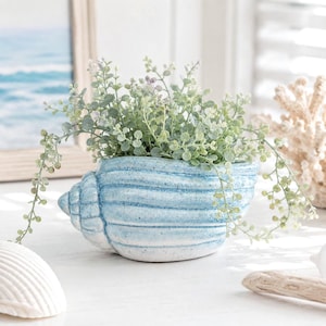Pode incluir: Um vaso de cerâmica azul-claro e branco em forma de concha, com um acabamento texturizado e salpicado. O vaso está preenchido com uma planta verde pendente com pequenas folhas redondas que caem sobre os lados do recipiente em forma de concha.