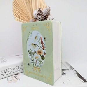 Könnte beinhalten: Eine hellgrüne, keramikbuchförmige Vase mit den Worten "BEES BOTANICALS" und einer Blumen- und Bienenillustration. Die Vase ist mit Trockenblumen und einem fächerförmigen Palmblatt gefüllt.