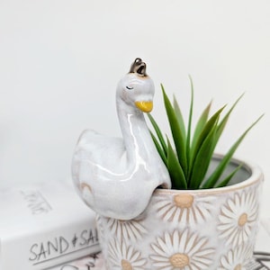 Op de afbeelding: Witte keramische zwanenplantenpot met een gele snavel en een kroon. De zwaan houdt een kleine witte plantenpot met een madeliefjespatroon en een groene succulent.