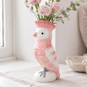 May include: A ceramic vase shaped like a cockatoo bird with a light pink and white glazed finish. The bird features a prominent crest, a small beak, and dark eyes. It stands on a white base with grey feet, holding a bouquet of pink roses and greenery.