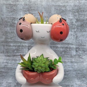 May include: A quirky white ceramic planter in the form of a person, holding a small coral-coloured pot filled with succulents. The planter has ladybird-shaped accents in pink, red, and tan. The face has closed eyes and a smiling mouth.