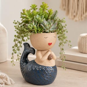 May include: A ceramic planter shaped like a mermaid with a beige head and a dark blue tail. The mermaid has closed eyes and a small red heart-shaped mouth. A variety of green succulents are planted in the top, spilling over the sides.