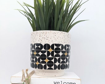 May include: A white ceramic planter with a black and gold geometric pattern. The planter is filled with green artificial grass and is sitting on a stack of white books with the text "welcome to" on the top book.