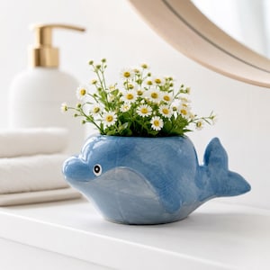 Könnte beinhalten: Ein glänzender, blauer Keramikübertopf in Delfinform, gefüllt mit einem Strauß kleiner weißer Gänseblümchen mit gelben Mitten. Die Delfinfigur hat einen glatten, abgerundeten Körper, eine ausgeprägte Schwanzflosse und ein aufgemaltes Auge.