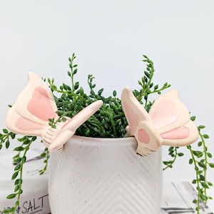 May include: Two ceramic butterfly plant stakes with light pink wings and a darker pink heart-shaped design. The butterflies are placed on a white ceramic pot filled with green plants. The pot has a textured surface.