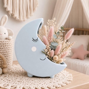 May include: A light blue ceramic crescent moon vase featuring a sleepy face with closed eyes, long eyelashes, and a pink cheek. The vase is filled with a decorative arrangement of dried beige grasses, pink bunny tails, and small green eucalyptus leaves.