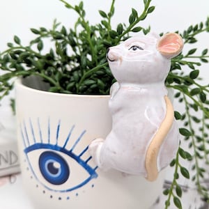 May include: White ceramic mouse figurine with a pink ear and a beige tail, climbing up the side of a white ceramic planter with a blue evil eye design.