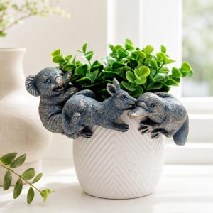 May include: A white ceramic planter with a chevron pattern featuring three dark bronze-toned animal figures attached to the rim: a koala, a kangaroo, and a platypus. The planter is filled with a small, vibrant green artificial plant.