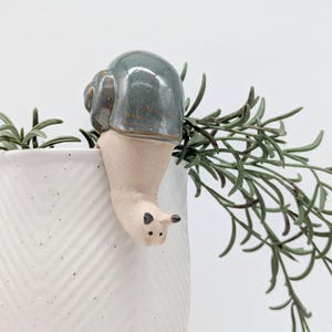 May include: A ceramic snail planter with a light blue and green glaze. The snail is hanging over the edge of a white planter with a chevron pattern. The snail has a small, black eye and a brown mouth.