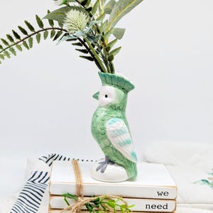 May include: A green ceramic parrot-shaped vase with greenery inside. The vase has a white eye and a crest. The bird is perched on a stack of white books tied with twine. The text "we need" is visible on the books.
