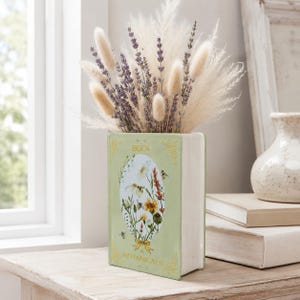 May include: A light green ceramic vase shaped like a book, featuring gold text that reads BEES & BOTANICALS and a central oval illustration of wildflowers and bees. The vase is filled with dried lavender sprigs and fluffy beige pampas grass.