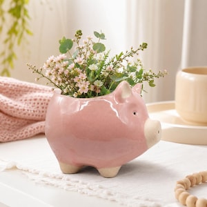 May include: A glossy pink ceramic pig-shaped planter featuring a smooth, rounded body, pointed ears, and a light beige snout and feet. The planter is filled with a bouquet of small, delicate pink flowers and green foliage.