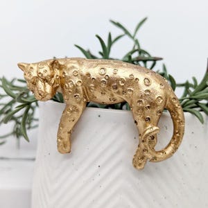 May include: A gold-painted figurine of a leopard, lying down with its head turned to the side. The figurine is detailed with spots and has a realistic appearance.