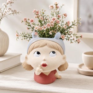 May include: A ceramic planter shaped like a head with a light blue headband, brown hair, and a red shirt. The figure has a finger held to its lips in a shushing gesture. The planter is filled with small, light pink flowers with green stems.