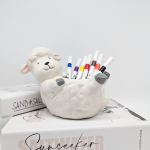 May include: White ceramic sheep figurine with a smiling face, holding a bunch of colourful markers in its belly. The sheep is sitting on a stack of books with the words "Sand & Salt" and "Sunseeker" visible.