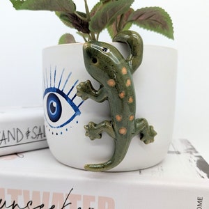 May include: White ceramic planter with a blue and white evil eye design and a green ceramic gecko figurine attached to the side. The planter is sitting on a stack of books.