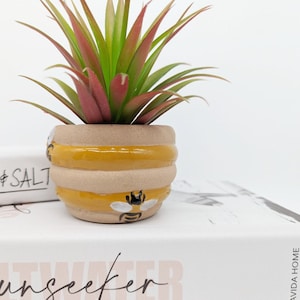 Op de afbeelding: Een kleine, gele en bruine keramische pot in de vorm van een honingraat met een bijendesign. De pot is gevuld met een groene en rode kunstmatige succulent.
