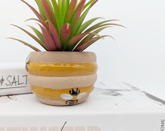 Op de afbeelding: Een kleine, gele en bruine keramische pot in de vorm van een honingraat met een bijendesign. De pot is gevuld met een groene en rode kunstmatige succulent.