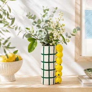 May include: A cylindrical white ceramic vase featuring a dark green grid pattern and a vertical handle composed of a stack of yellow lemons. The vase is filled with green eucalyptus branches and small white flowers, standing upright.