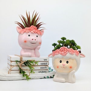 Peut inclure: Deux pots de fleurs en céramique en forme de visage d'animal. Le pot de cochon est rose avec une couronne de fleurs roses et le pot de mouton est blanc avec une couronne de fleurs roses. Les deux pots contiennent une plante succulente. Les pots sont posés sur une pile de livres blancs avec le texte "we need each other" écrit sur les livres.
