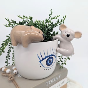 May include: A white ceramic planter with a blue evil eye design. A small ceramic koala figurine is climbing the side of the planter, and a small ceramic wombat figurine is perched on the rim.