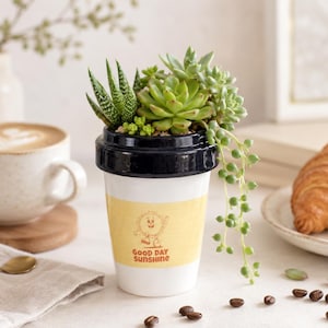 Puede incluir: Una maceta de cerámica blanca con forma de vaso de café desechable con tapa negra. El vaso presenta una banda amarilla con un gráfico de sol de dibujos animados y el texto "GOOD DAY SUNSHINE". Está lleno de varias suculentas verdes, incluida una planta rosario colgante.