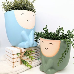 May include: Two ceramic planters shaped like smiling faces, one blue and one green, with plants growing out of the top. The blue planter is sitting on a stack of books with the text "we need each other" on the top book.